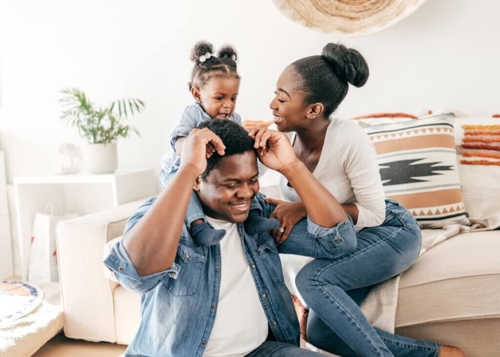 a set of parents with their young child who is sitting on the dads back in their living room. thinking about their estate plan for their daughter