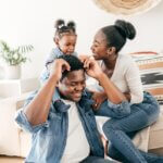 a set of parents with their young child who is sitting on the dads back in their living room. thinking about their estate plan for their daughter