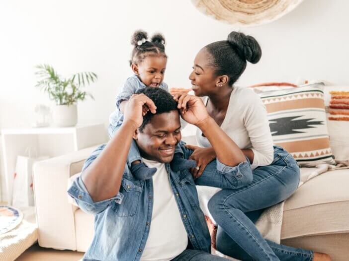 a set of parents with their young child who is sitting on the dads back in their living room. thinking about their estate plan for their daughter