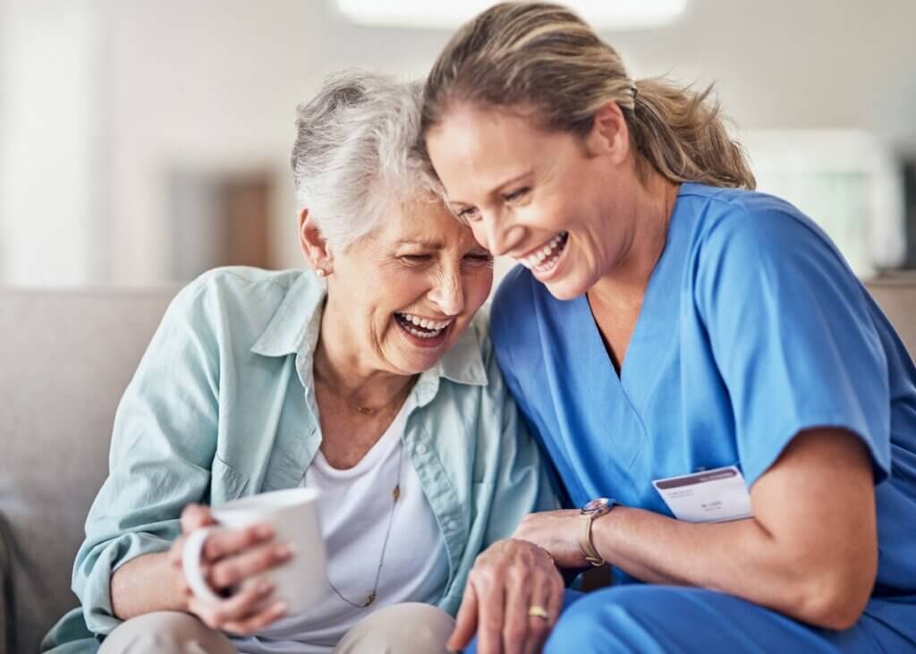 the caregiver and the patient are on the couch laughing with one another and sitting right next to each other. the caregiver is taking very good care of her patient.