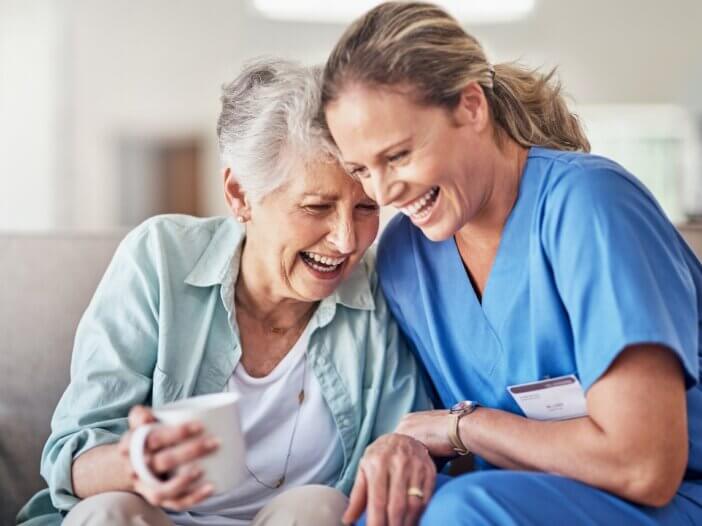 the caregiver and the patient are on the couch laughing with one another and sitting right next to each other. the caregiver is taking very good care of her patient.