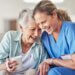 the caregiver and the patient are on the couch laughing with one another and sitting right next to each other. the caregiver is taking very good care of her patient.