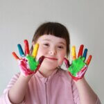 a little girl with special needs, down syndrome with her hands painted different colors, showing them to the camera while she smiles.