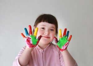 a little girl with special needs, down syndrome with her hands painted different colors, showing them to the camera while she smiles.