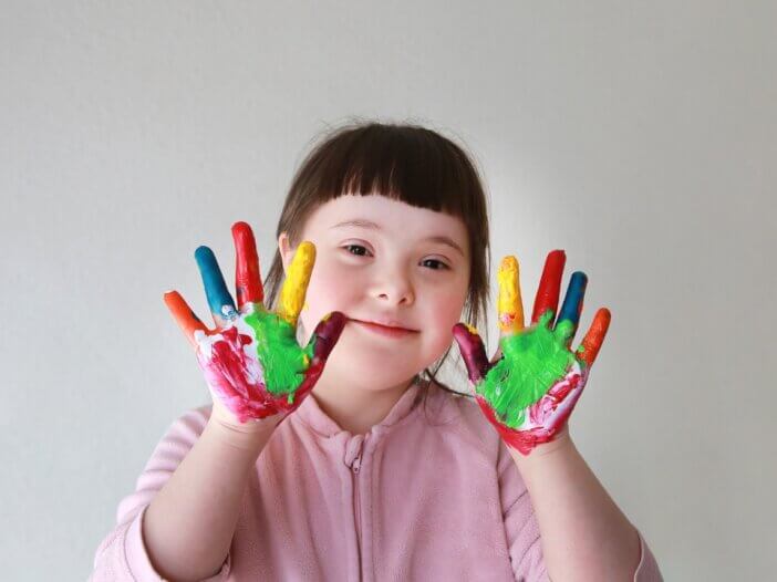 a little girl with special needs, down syndrome with her hands painted different colors, showing them to the camera while she smiles.