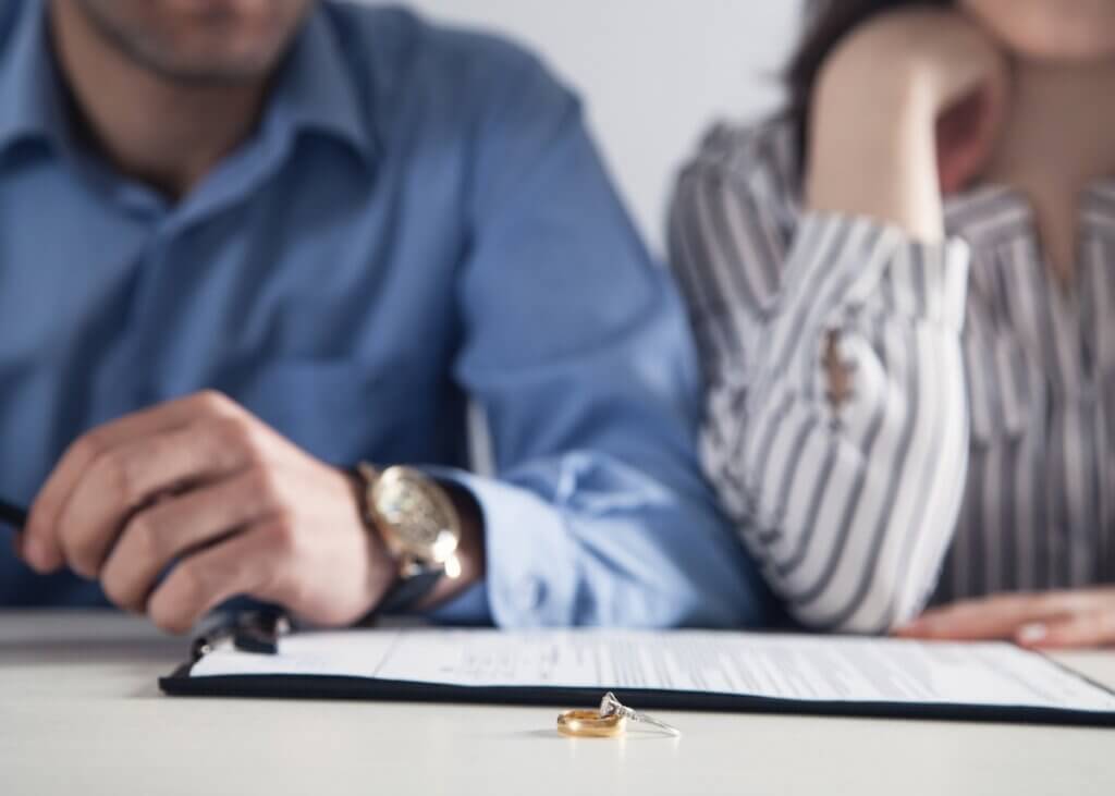 a picture of two wedding rings- one golden band and one diamond band and there is a couple in the background with divorce paperwork.