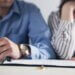 a picture of two wedding rings- one golden band and one diamond band and there is a couple in the background with divorce paperwork.