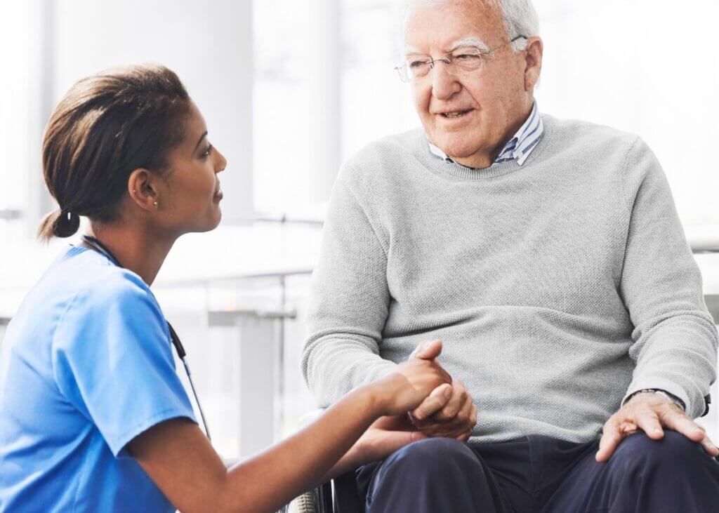 Doctor kneeling and holding the hand of a man in a wheelchair, offering support and care.