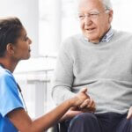 Doctor kneeling and holding the hand of a man in a wheelchair, offering support and care.