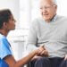 Doctor kneeling and holding the hand of a man in a wheelchair, offering support and care.