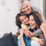 a grandma, a daughter, and granddaughter all hugging- showing the three generations and they are all laughing and smiling.