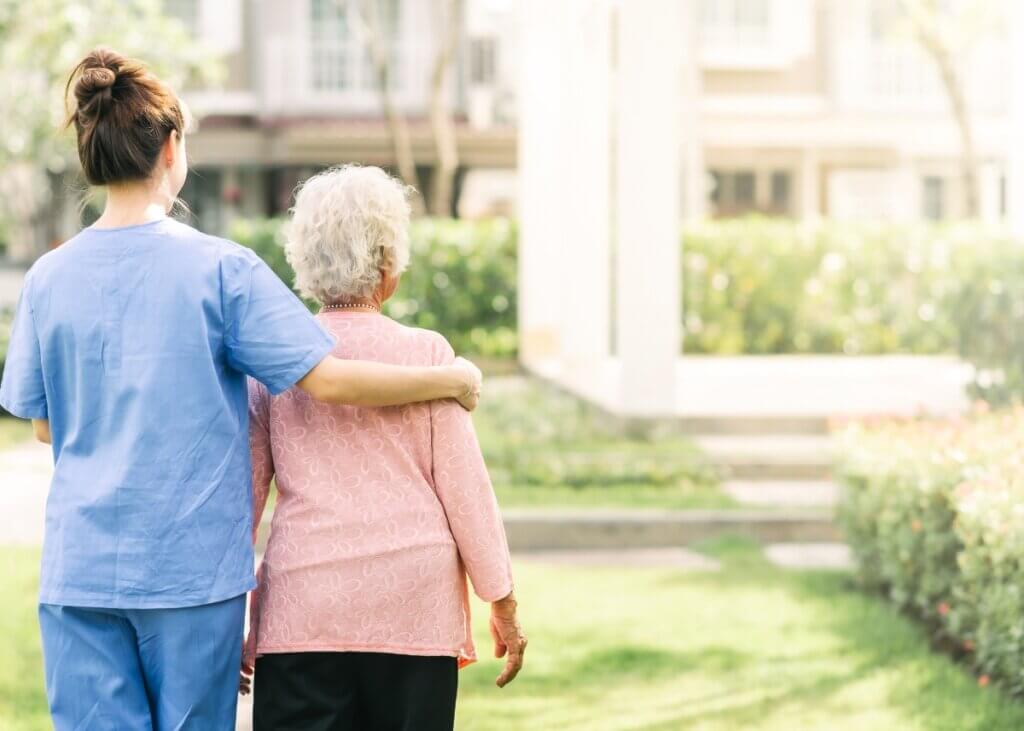 a caregiver and her patient with their backs facing us. the caregiver has her arm around her patient while they are on a walk.