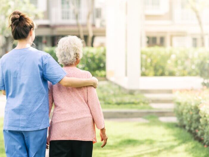 a caregiver and her patient with their backs facing us. the caregiver has her arm around her patient while they are on a walk.