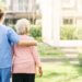 a caregiver and her patient with their backs facing us. the caregiver has her arm around her patient while they are on a walk.