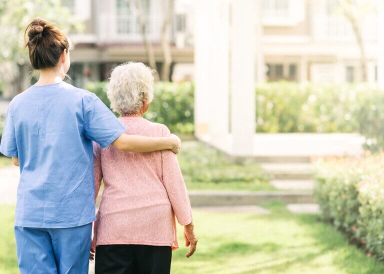 a caregiver and her patient with their backs facing us. the caregiver has her arm around her patient while they are on a walk.