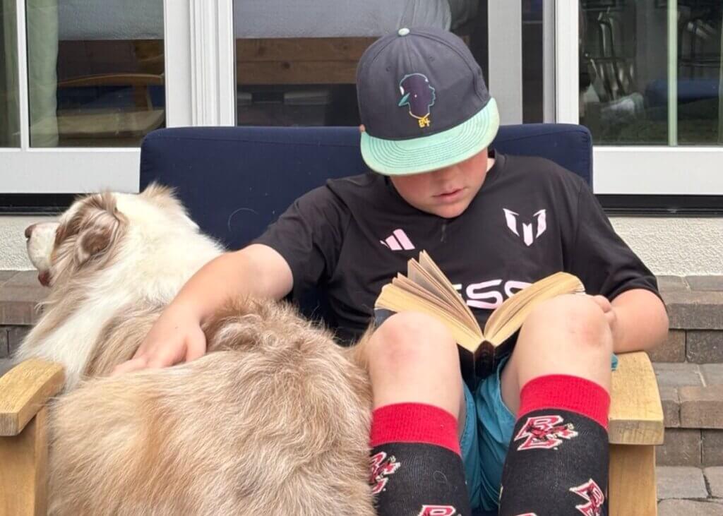 a kid with reading a book on the chair, petting his dog sitting next to him.