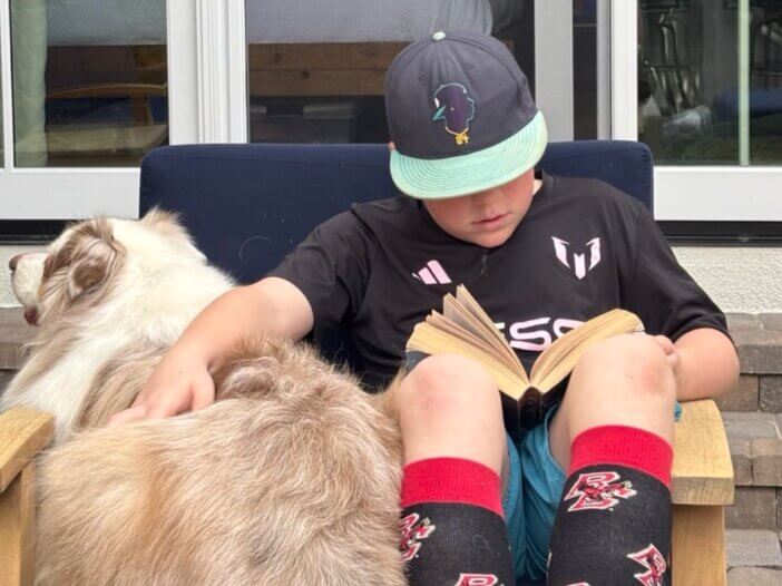 a kid with reading a book on the chair, petting his dog sitting next to him.