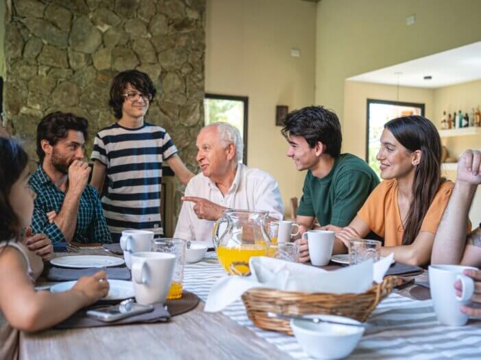 family having breakfast at the dinning room table, all in harmony and discussing a serious manner. the grandpa is talking and everyone is sitting around him listening.