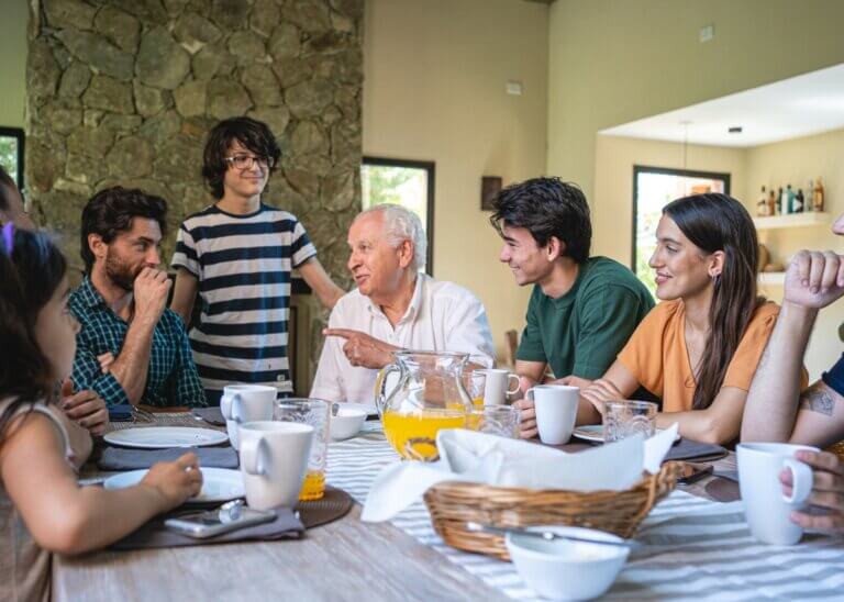 family having breakfast at the dinning room table, all in harmony and discussing a serious manner. the grandpa is talking and everyone is sitting around him listening.