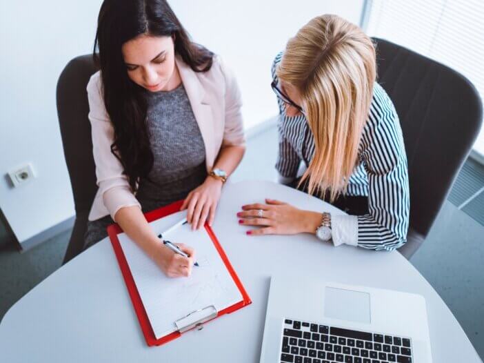 overhead shot of two women in a meeting. one of them is writing things down on a piece of paper while the other one is advising.