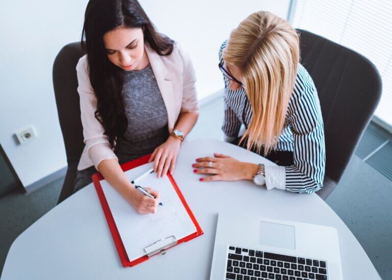 overhead shot of two women in a meeting. one of them is writing things down on a piece of paper while the other one is advising.