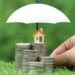 a little house on top of a stack of coins and an umbrella protecting these coins and house