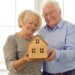 an older couple smiling together holding a small wooden house, symbolizing their trusts and assets.