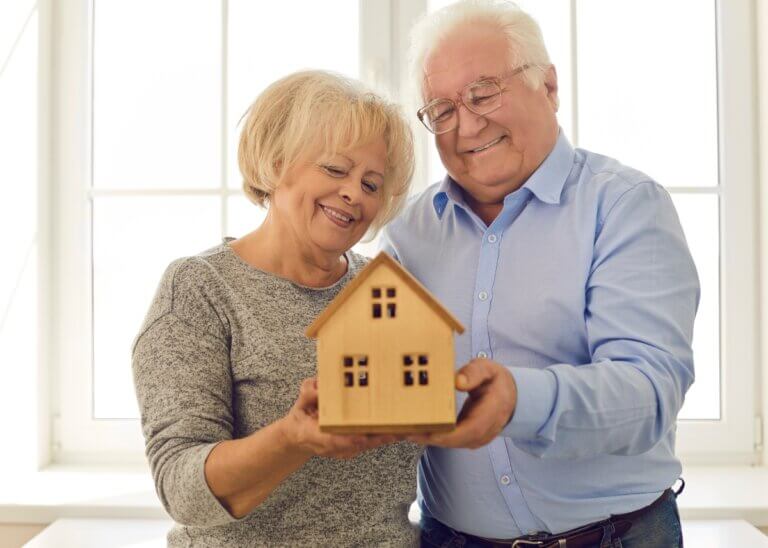 an older couple smiling together holding a small wooden house, symbolizing their trusts and assets.