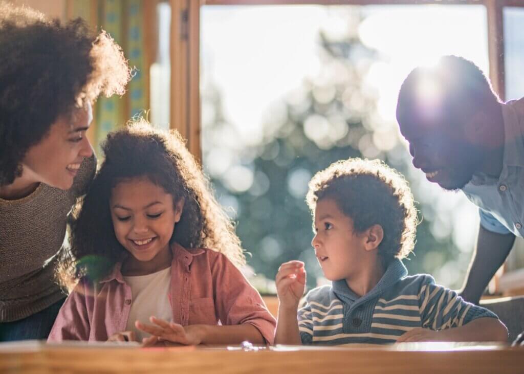 a younger couple with their two younger kids. they are sitting at the kitchen table all laughing together